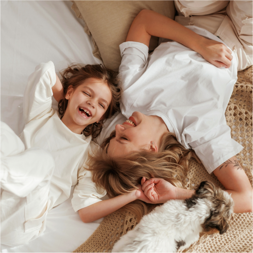 Anya és lánya nevetnek az ágyon, mellettük egy kis kutya